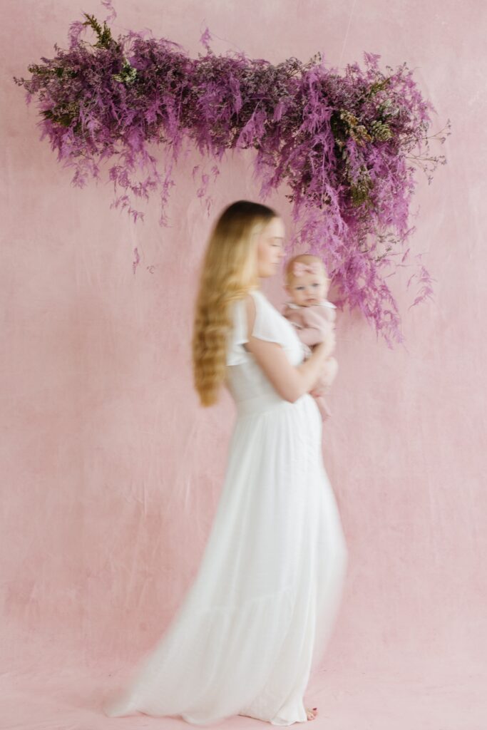 Mother holding baby on a pink painted backdrop and  purple floral installation. Utah Motherhood Photographer Mary Horne Nelson